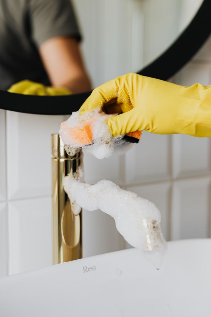 Cleaner in yellow gloves scrubbing a bathroom faucet with soap suds